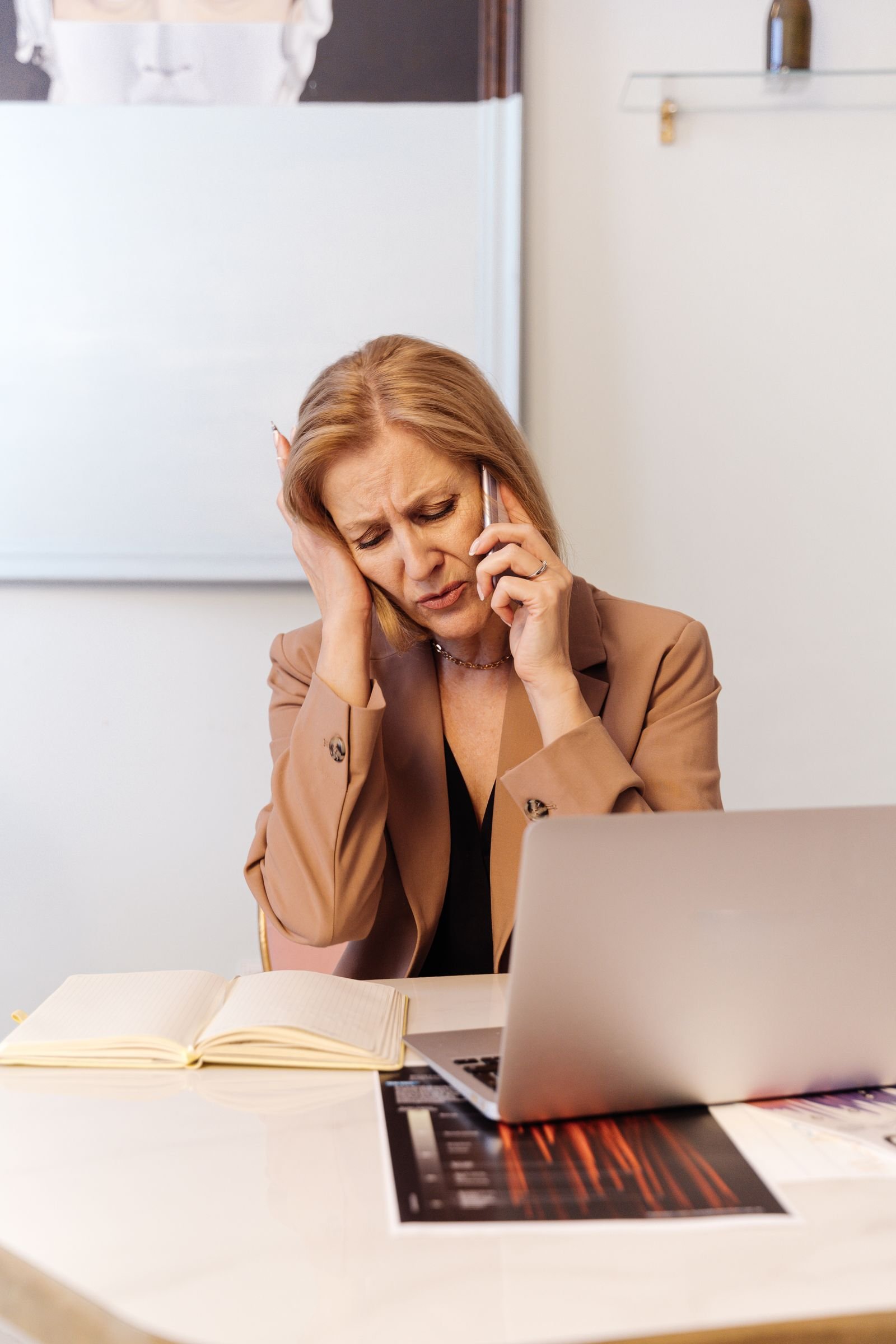 A businesswoman sits at her desk on the phone, looking concerned while checking her laptop in a bright office