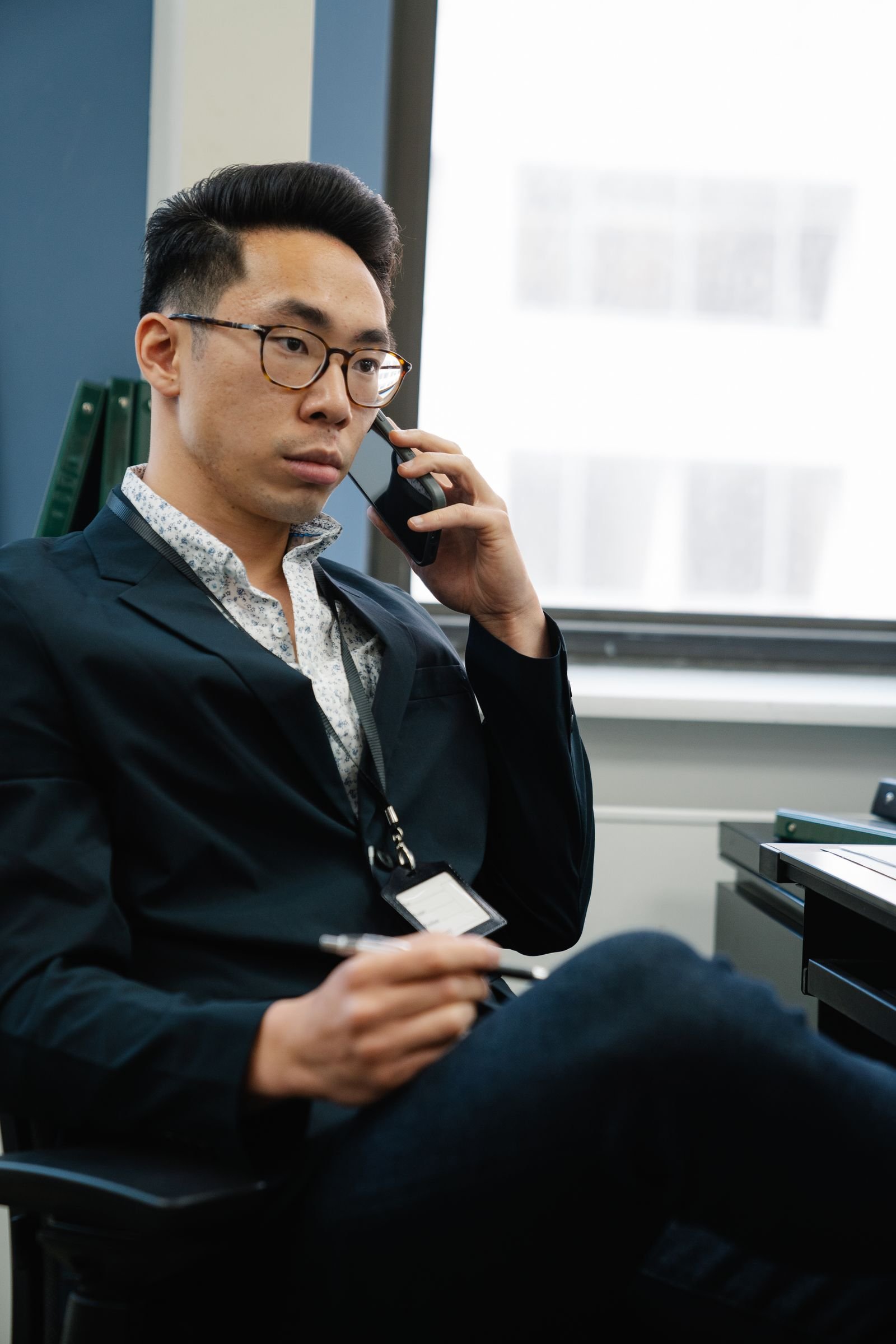 A businessman in an office holds a pen and takes a serious phone call at his desk
