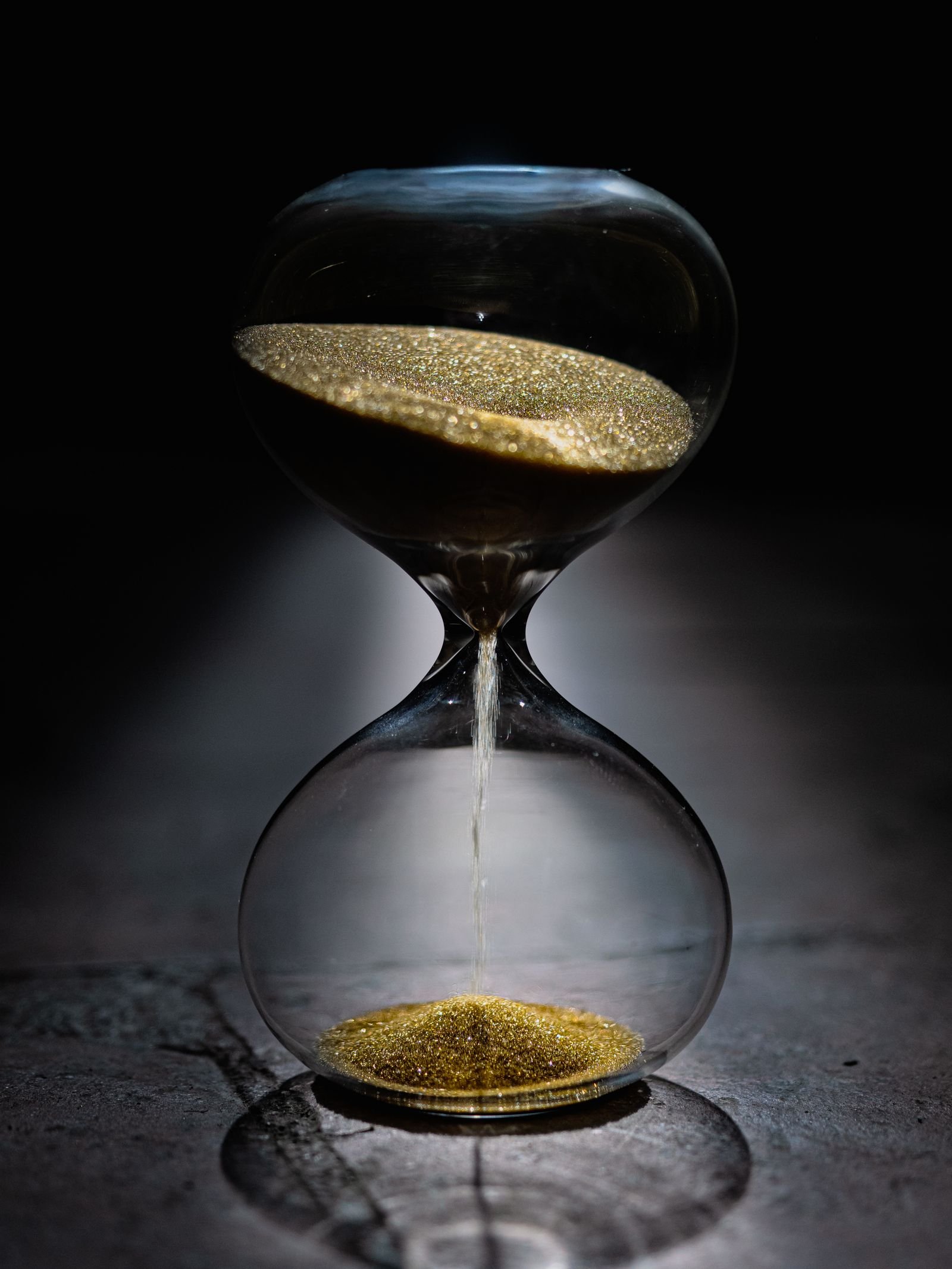 An hourglass with gold sand flows on a dark reflective surface